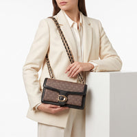 A model in a cream blazer and white shirt holds a brown monogram shoulder bag with a black leather trim and chain strap featuring gold links, set against a clean white background.