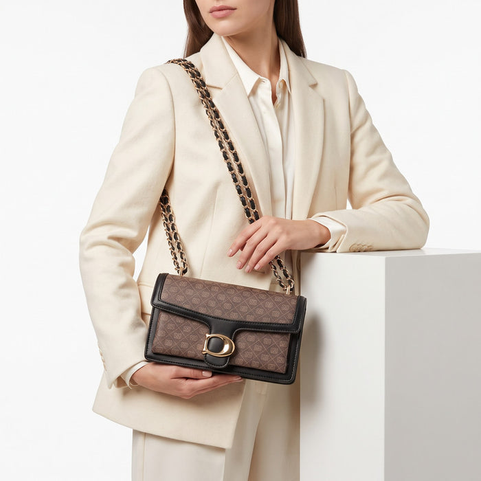 A model in a cream blazer and white shirt holds a brown monogram shoulder bag with a black leather trim and chain strap featuring gold links, set against a clean white background.