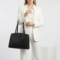 A model in a white blazer and trousers holds a black leather tote bag with gold hardware. The bag features the brand name 'BURGA' in gold on the front and a hanging leather tag.