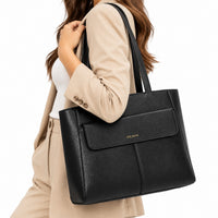 A woman wearing a beige blazer and white top carries a large black leather tote bag with a front flap pocket. The bag has a small gold 'DEJAYU' logo on the front and is photographed against a clean white background.